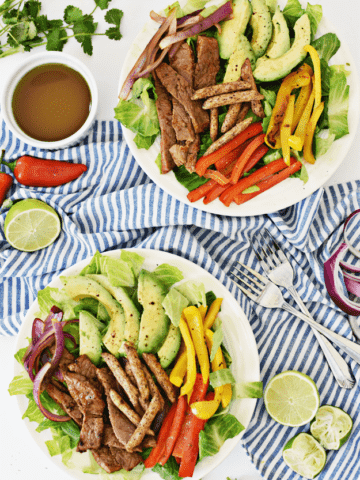 Overhead shot of two white plates steak, avocados, peppers, and salad on white and blue napkin.