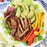 Overhead shot of large plate of steak, avocado, peppers, and onion on white and blue napkin.