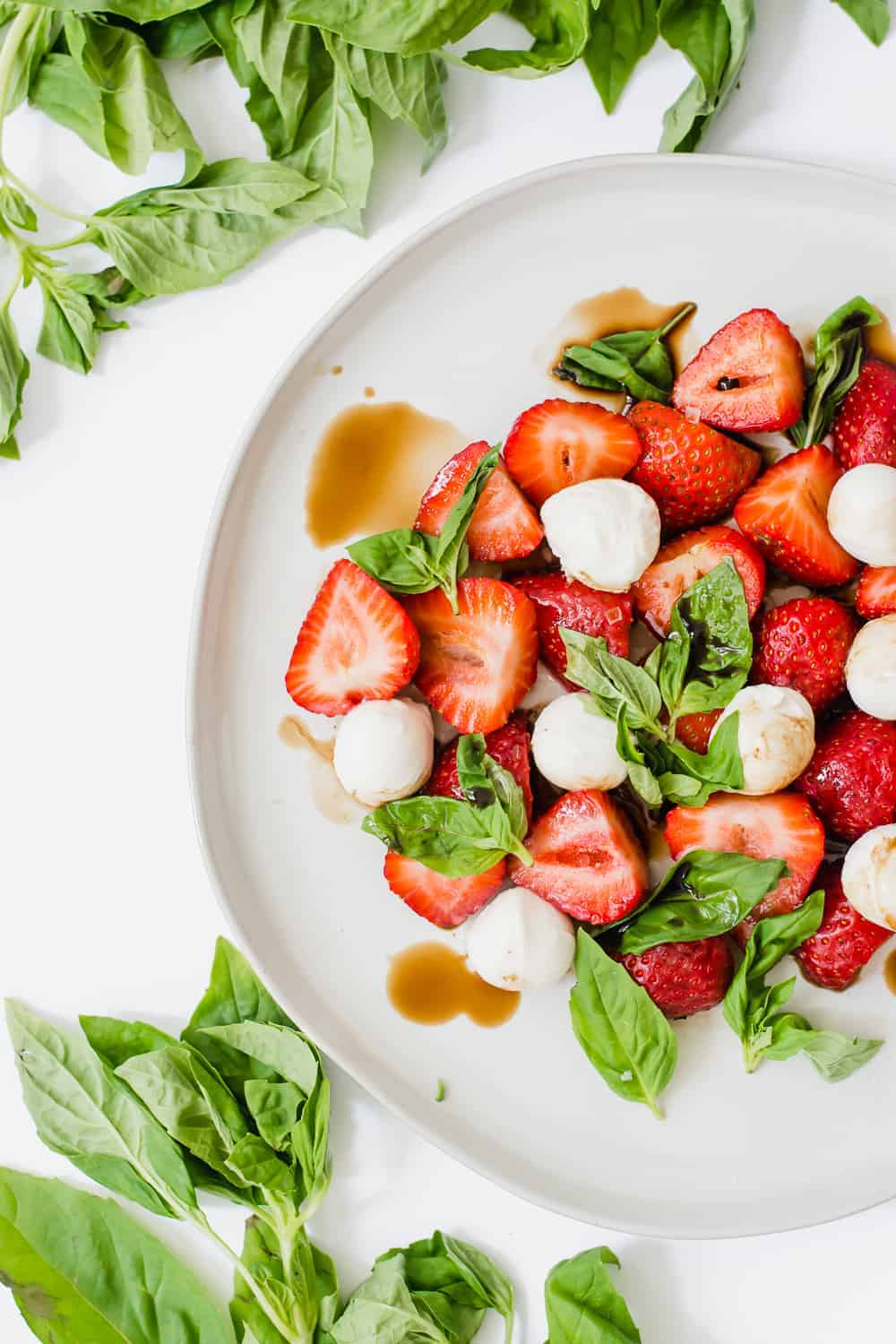 Overhead shot of halve strawberries on white plate with basil leaves and mozzarella balls.