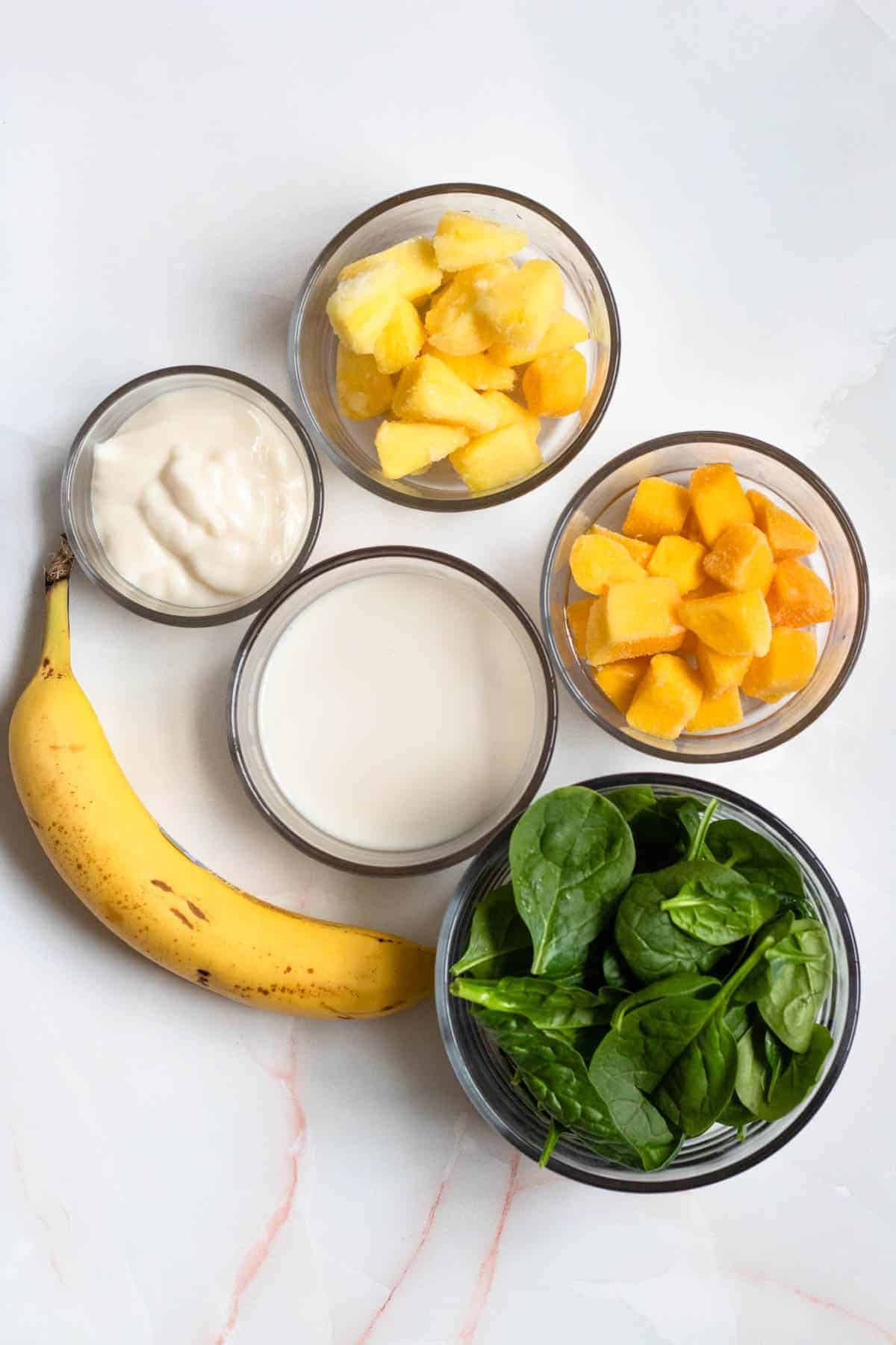 bowls of fruit, banana, spinach, almond milk, and yogurt on a light gray background