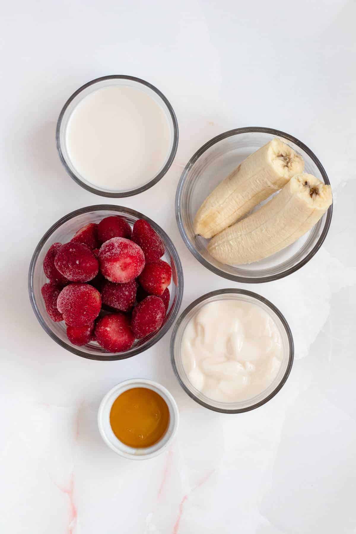 Small glass bowls with recipe ingredients; Frozen Strawberries, Banana, Almond Milk, Yogurt, and Honey on a white background