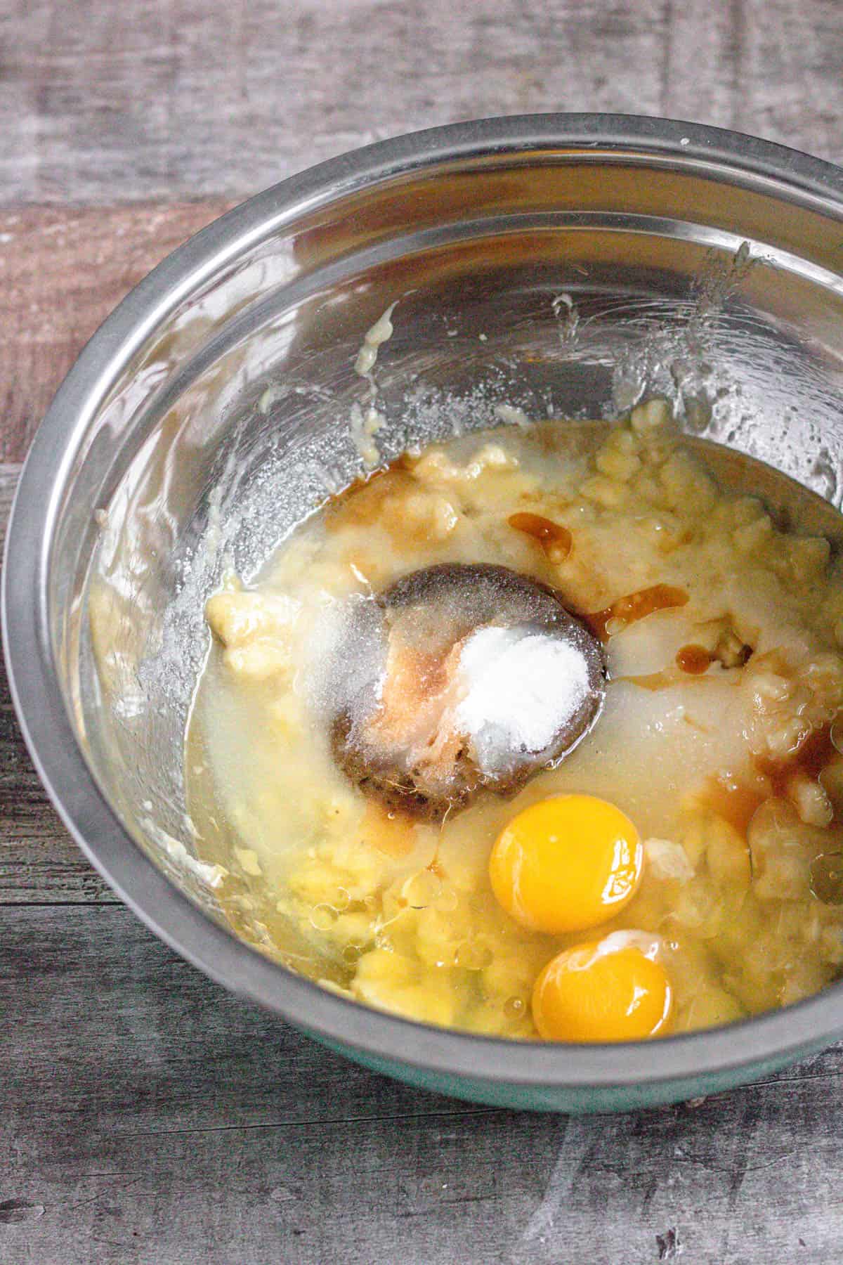 mashed bananas, brown sugar, granulated sugar, vegetable oil, eggs, and vanilla in a metal bowl with a dark colored background