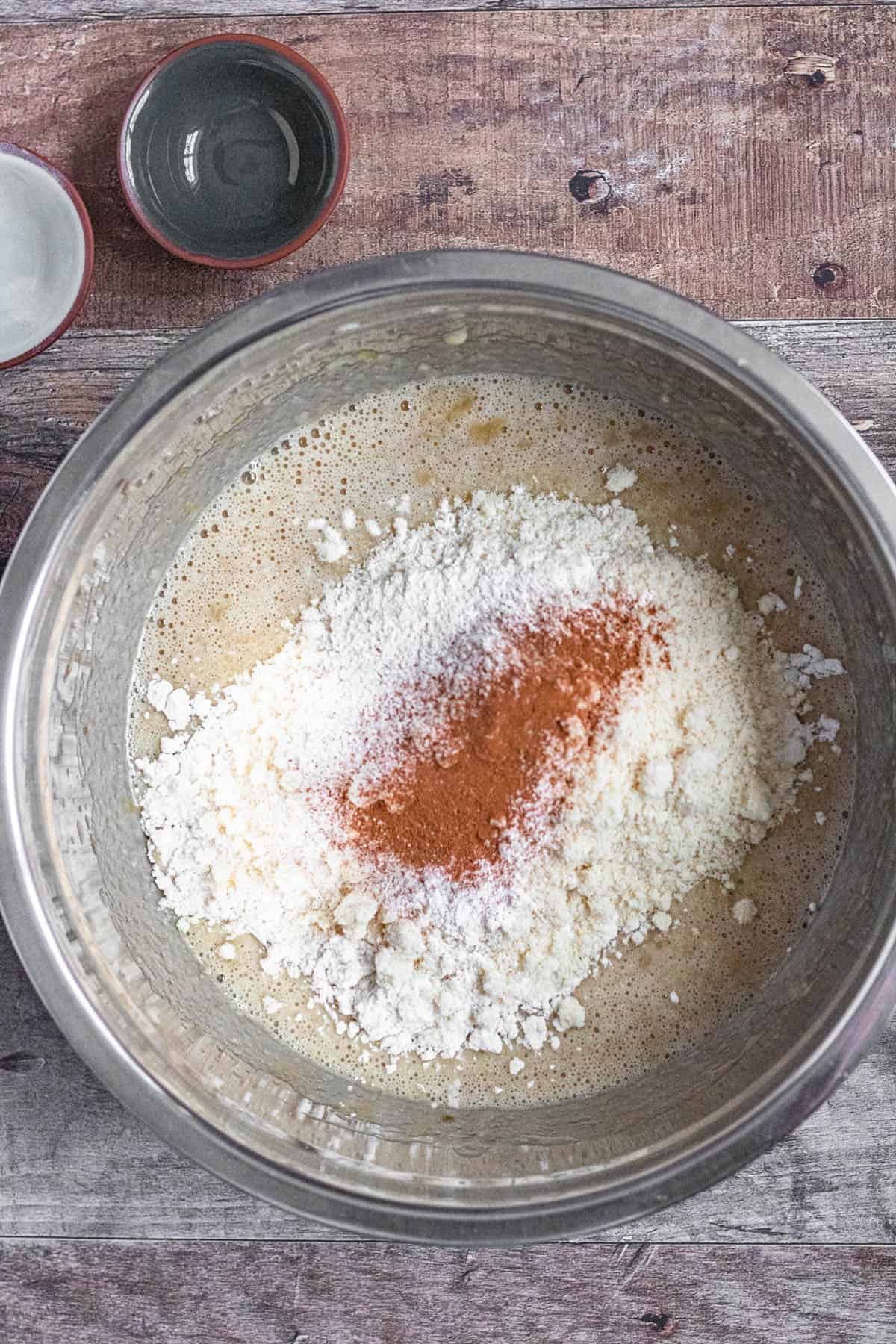 gluten free flour, almond flour, baking powder, baking soda, salt, and cinnamon in metal bowl with mixed wet ingredients and mashed bananas with a dark colored background