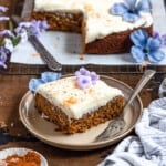 a slice of gluten free carrot cake with the rest of the cake behind it on a dark colored background