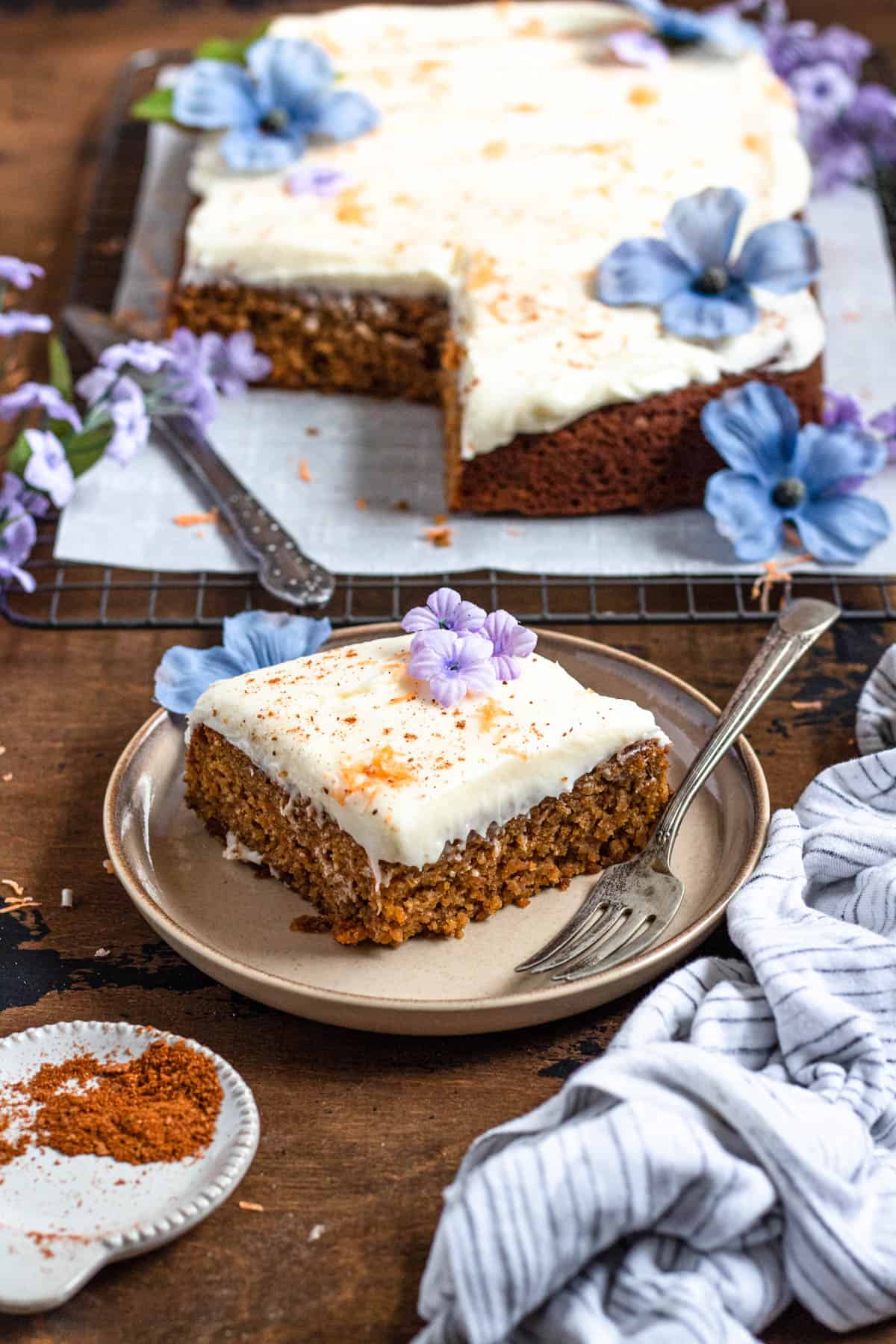 a slice of gluten free carrot cake on a round light colored plate with a silver fork, purple flowers topping the cake, and a striped towel lying next to the plate