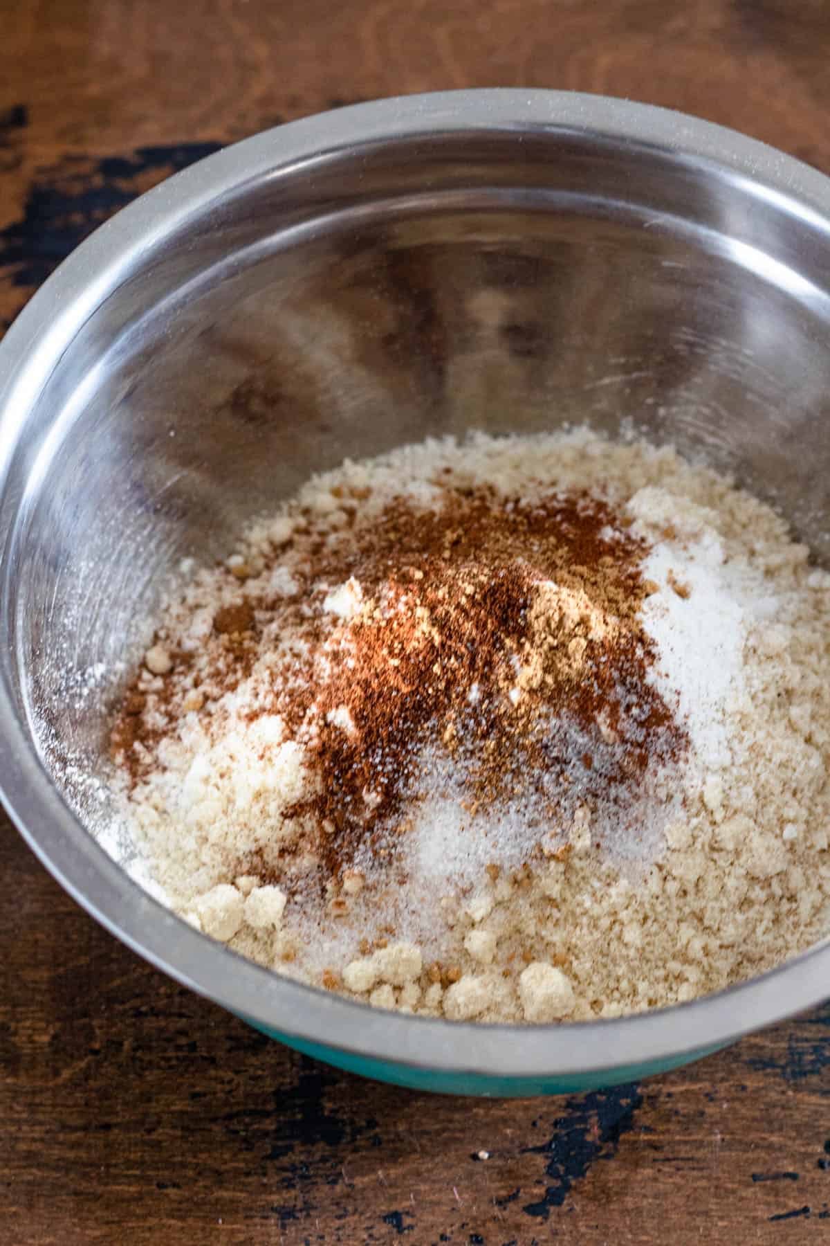 metal bowl with dry ingredients; gluten free flour, almond flour, baking powder, baking soda, cinnamon, ginger, nutmeg, cloves, and salt with a dark wood colored background