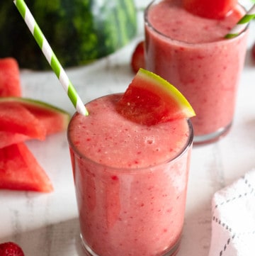 two glasses filled with watermelon smoothie with a green and white striped straw in front of a light backdrop