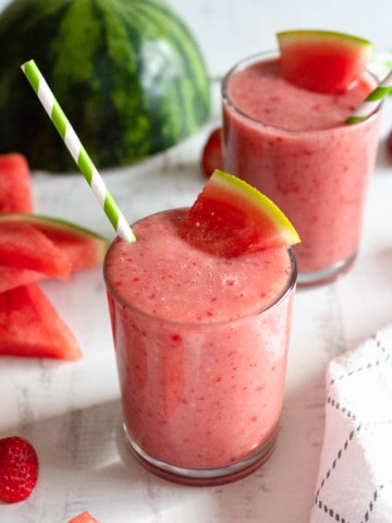 two glasses filled with watermelon smoothie with a green and white striped straw in front of a light backdrop