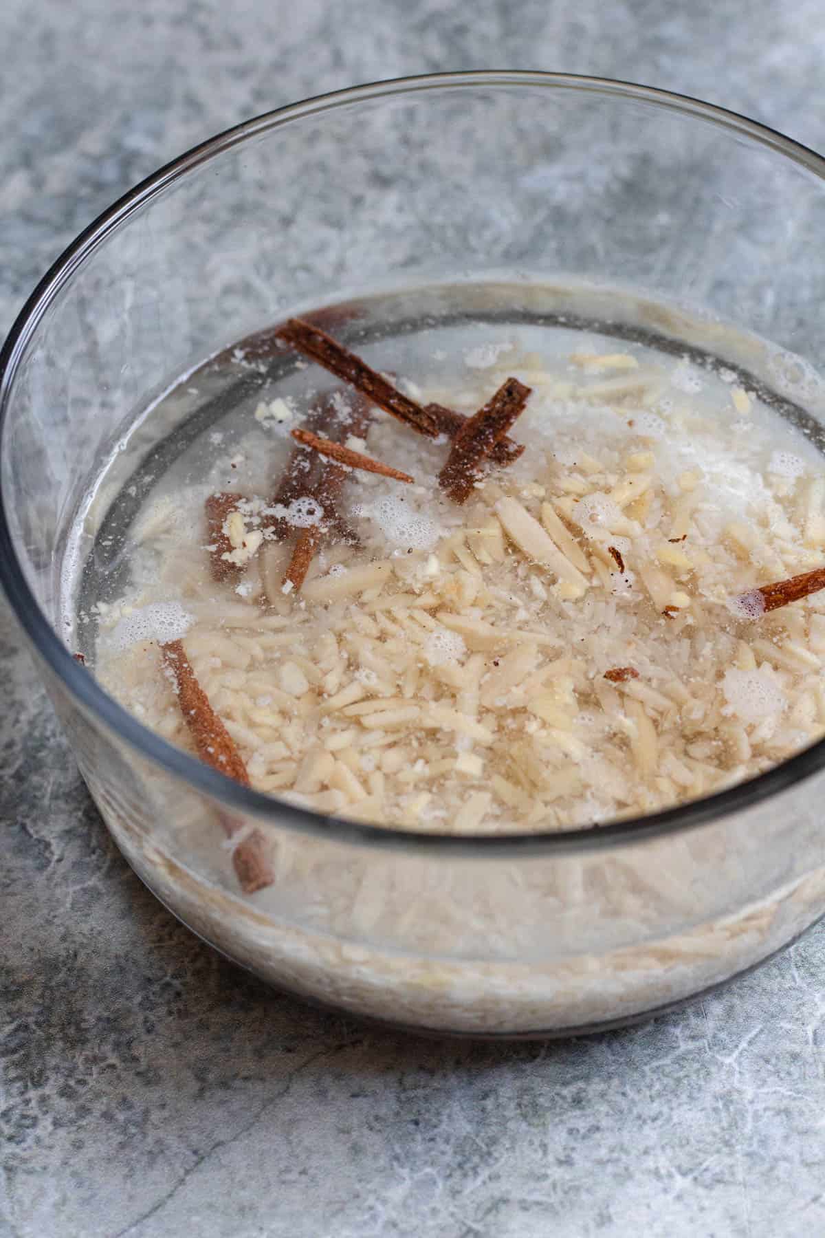 glass bowl with water, rice, slivered almonds, and cinnamon sticks on a light colored background