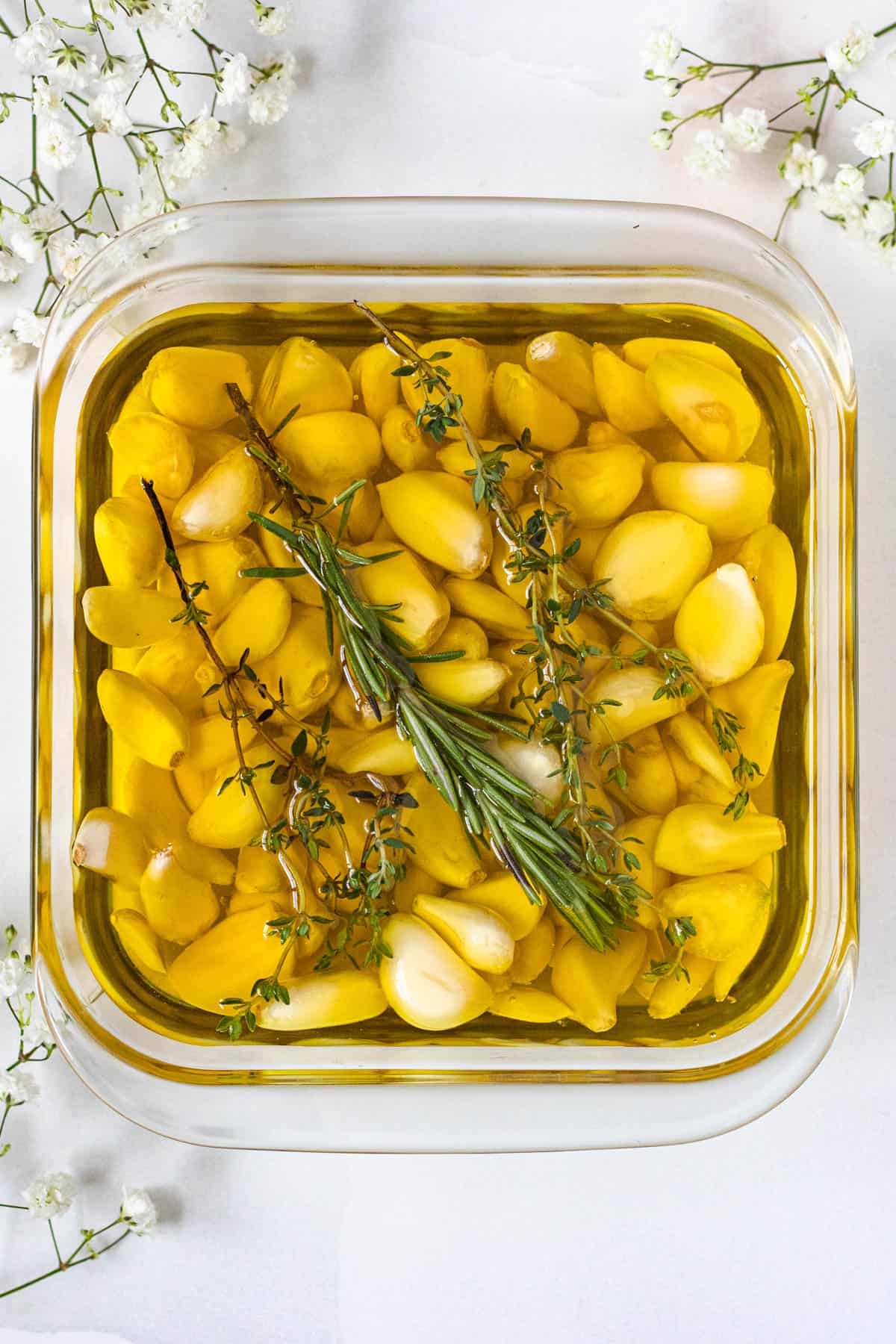 garlic cloves with olive oil, rosemary, and thyme in an oven safe pan with a light colored background