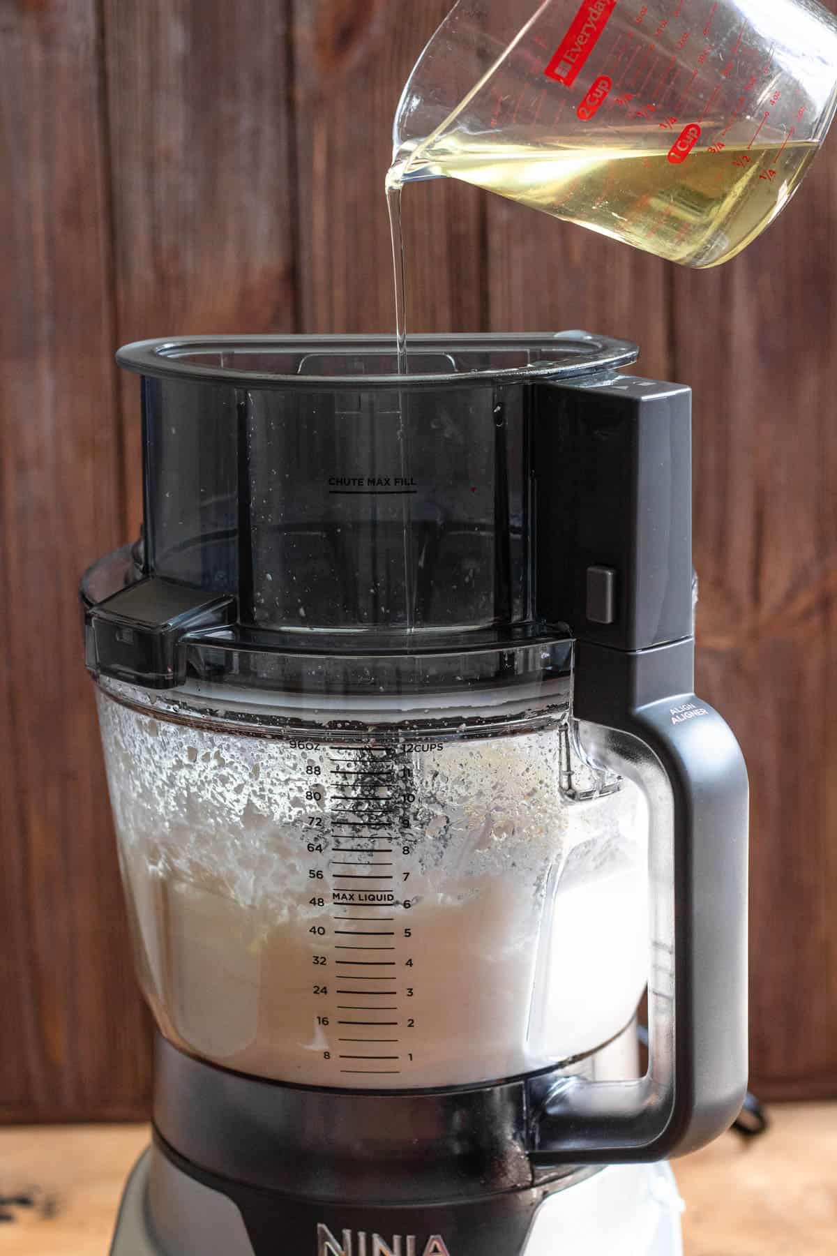food processor with blended garlic cloves, salt, and lemon juice with canola oil slowly being poured in through the lid with a dark colored background