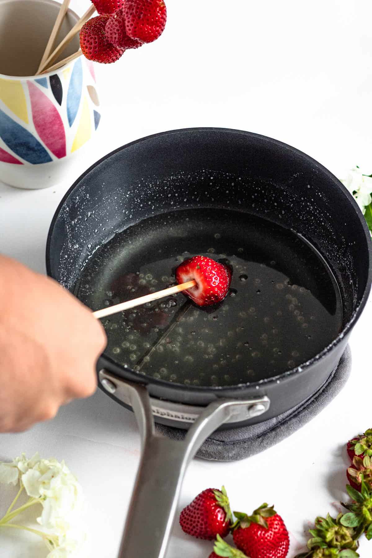 bamboo skewer with strawberry being dipped in water and sugar mixture in a pot with a light colored background